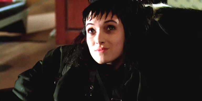 Winona Ryder as Lydia Deetz in Beetlejuice sitting in a chair all dressed in black with a pale face and jagged bangs, looking up sort of smugly.