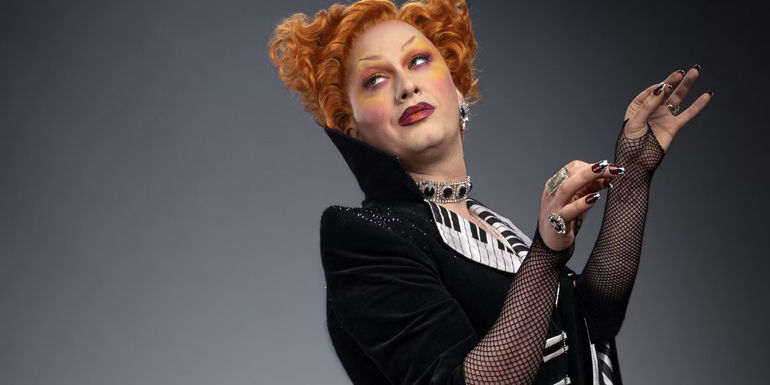 In this promo photo, Jinkx Monsoon's unnamed character in Doctor Who holds their hands up like they're conducting.