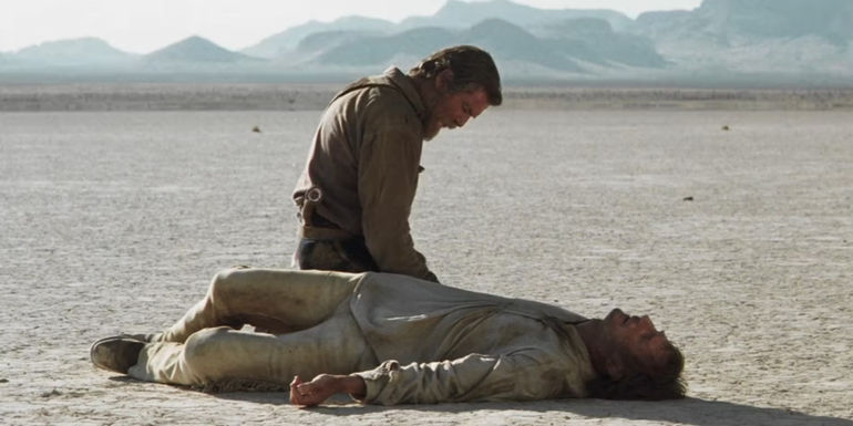 Gideon (Pierce Brosnan) kneeling over a prone Carver (Liam Neeson) after shooting him in the desert in Seraphim Falls