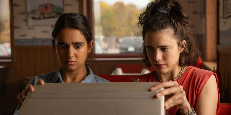 Geraldine Viswanathan and Margaret Qualley as Marian and Jamie looking shocked while opening a briefcase in Drive-Away Dolls