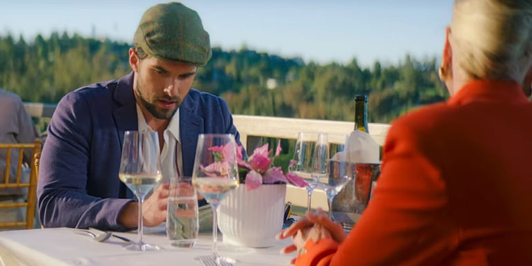 Carter looks down while sitting at an outside table with Margo in Holiday in the Vineyards