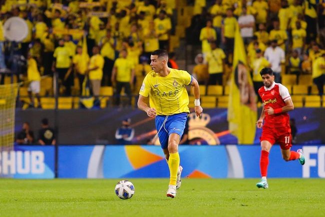 Cristiano Ronaldo in a yellow kit dribbles a soccer ball with an opponent in red behind him on a pitch, with spectators in the background.