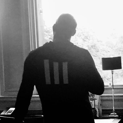 Vin Diesel's silhouette by a window with trees, near a lamp and desk, donning a striped jacket.
