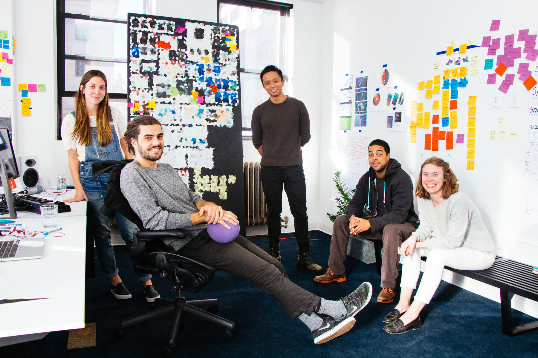 A group of people smiling at the camera, with sticky notes and drawings on the walls behind them
