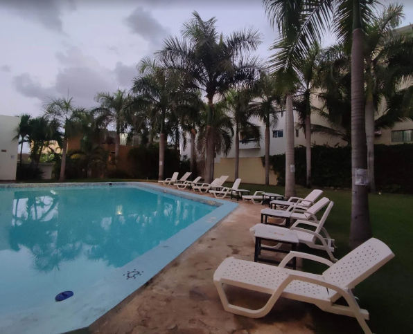 Patio interior de un hotel con piscina turquesa en primer plano, rodeado de vegetación tropical y una hamaca