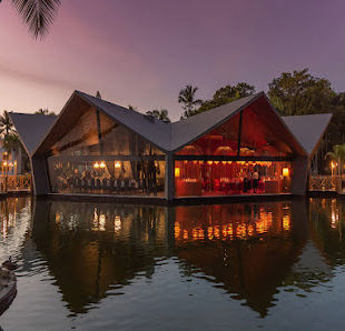 Elegant restaurant with glass walls and a zigzag roof situated over a lake, reflecting its warm interior lighting during a tropical sunset."