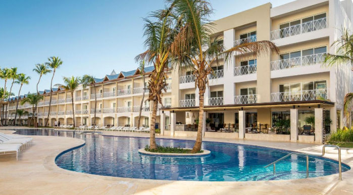 "Vista exterior de un resort tropical con una gran piscina, palmeras y sillas de playa frente a un edificio de habitaciones con balcones bajo un cielo azul."