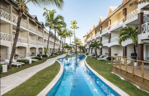 A long, winding blue swimming pool running through a tropical resort. The pool is flanked by green grass, white lounge chairs, tall palm trees, and symmetrical three-story hotel buildings with white balcony railings.