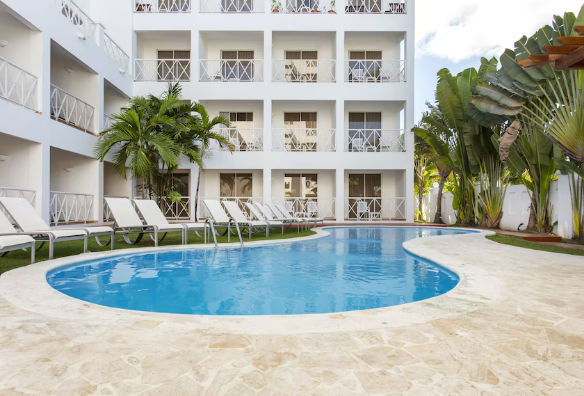 "Blue water swimming pool surrounded by white sun loungers in front of a white hotel building with balconies and tropical palm trees."