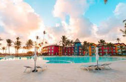 Large central pool including a colorful children's water park, surrounded by dense palm trees, bright yellow buildings with thatched roofs and, in the background, a white sand beach and the turquoise ocean.