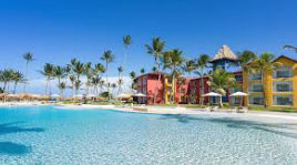 "Tropical beachfront resort with a large blue pool, palm trees, colorful buildings and a beach in the background."