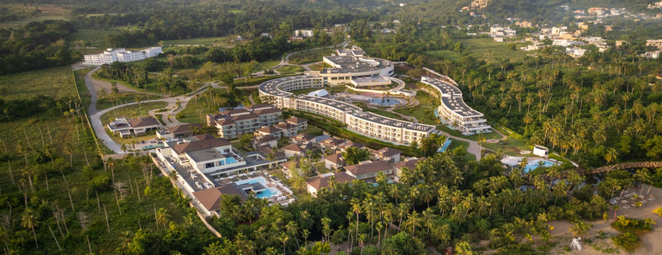 "Aerial view of a large hotel complex surrounded by dense tropical vegetation and palm trees. Several white buildings with curved and linear designs stand out, along with a central pool area and direct access to the beach sand in the lower right corner."