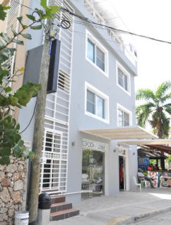 "Vista exterior de un edificio de hotel de varios pisos en color gris con marcos blancos. En la planta baja se observa la entrada de cristal a la recepción y un pequeño comercio adyacente."