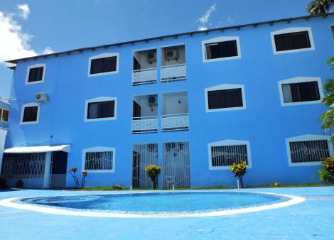 Blue hotel exterior with outdoor pool, balconies and green areas on a sunny day.