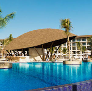 Pool bar with palapa roof at a luxury resort, with large blue pool and palm trees.
