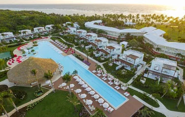 Aerial view of a coastal resort at sunset, highlighting a very long pool, a large palapa, white buildings and the ocean in the background.