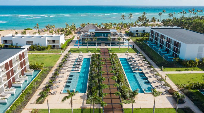Vista aérea de drones del Live Aqua Beach Resort Punta Cana, que muestra modernos edificios de hotel de varios pisos con balcones que flanquean dos largas piscinas turquesas y pasarelas ajardinadas. El complejo se extiende hasta una playa de arena blanca 