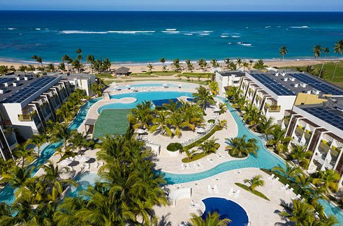 Aerial view of a luxury beachfront resort featuring large winding swimming pools, palm trees, and modern white buildings with solar panels, overlooking the turquoise ocean and sandy beach.