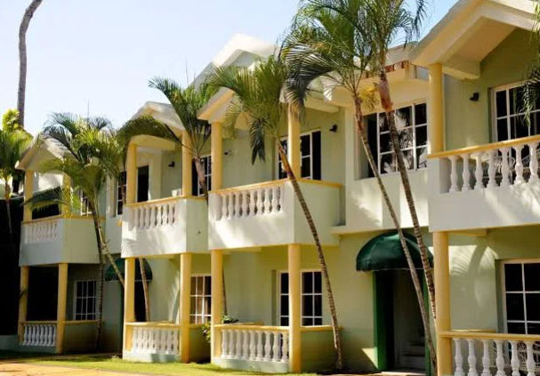 "Exterior facade of a colonial-style hotel in Punta Cana, with two-level buildings, walls in light green and yellow tones, balconies with white balustrades and tall palm trees in the foreground."
