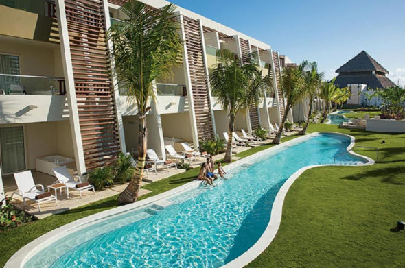 Vista aérea de un resort de lujo frente al mar que muestra grandes piscinas serpenteantes, palmeras y modernos edificios blancos con paneles solares, con vista al océano turquesa y la playa de arena.