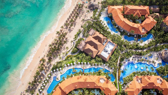 Aerial view of the Majestic Elegance Punta Cana resort showing the white sand beach, turquoise sea, extensive blue swimming pools, and buildings with terracotta roofs.