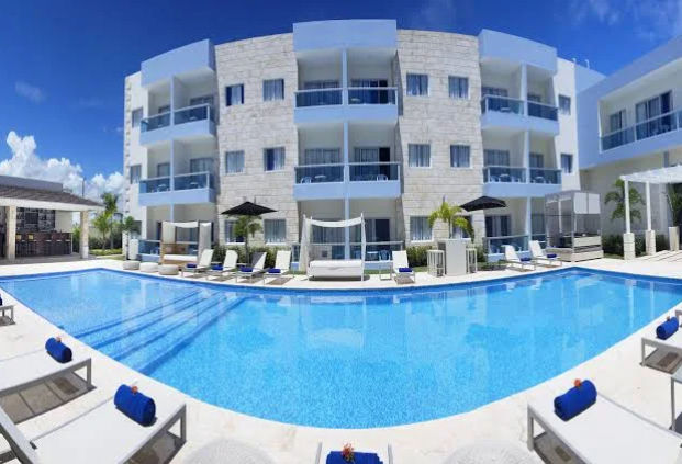 Pool area of ​​a modern hotel with sun loungers, cabanas and a four-story building in the background under a cloudy blue sky.