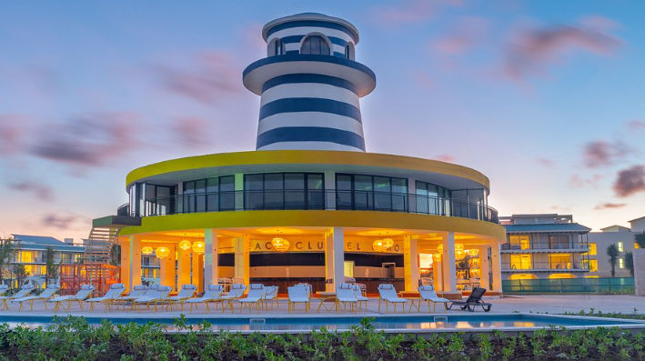 "Blue and white striped decorative lighthouse over the illuminated Beach Club at dusk, with lounge chairs by the pool in the foreground."