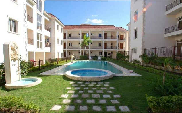 Interior courtyard of a residential complex with green lawn, a rectangular swimming pool, a circular jacuzzi in the foreground and white Mediterranean-style apartment buildings around it.