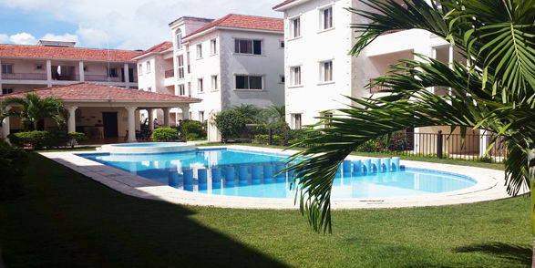 Patio interior de un complejo residencial con césped verde, una piscina rectangular, un jacuzzi circular y edificios de apartamentos blancos.