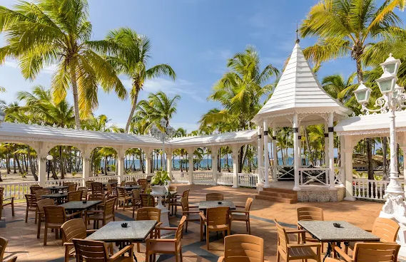 muestra una terraza al aire libre ubicada en un entorno tropical, caracterizada por una arquitectura de estilo colonial con estructuras blancas que incluyen una pérgola y un gazebo. El espacio está equipado con mesas y sillas distribuidas para los comensa
