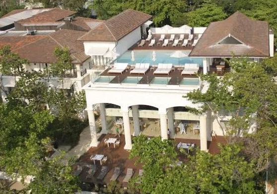 Presenta una vista aérea de un elegante complejo vacacional caracterizado por su arquitectura de estilo clásico, distinguida por columnas blancas y techos de teja en tonos marrón rojizo. La propiedad cuenta con una terraza superior equipada con una piscin