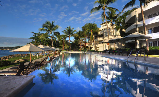 An outdoor infinity pool located by the sea, surrounded by tropical palm trees and umbrellas, with the structure of a hotel building in the background under a clear blue sky.