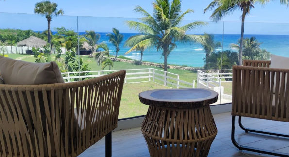 A través de una barandilla de cristal, se aprecia un área ajardinada con césped verde, palmeras y una estructura con techo de paja al fondo. El paisaje se completa con una vista despejada del mar Caribe, caracterizado por sus tonos turquesa y azul profund