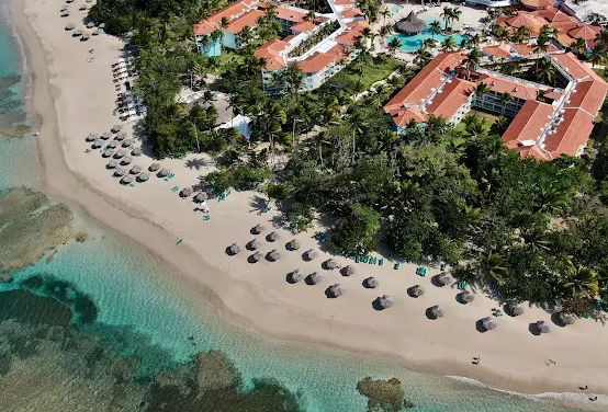 "Aerial view of a tropical resort next to a white sand beach with umbrellas and crystal clear waters."