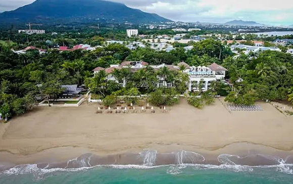"Aerial view of a beach resort with white buildings surrounded by vegetation, facing a sandy coast and turquoise sea."
