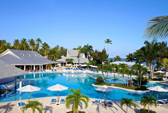 Vista panorámica de una piscina grande y cristalina en un resort tropical, rodeada de palmeras, sombrillas blancas y edificios con techos grises bajo un cielo despejado.