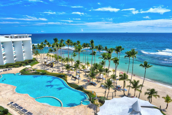 Vista panorámica de un complejo turístico tropical con una piscina azul turquesa donde una pareja camina por un borde divisorio, rodeada de sombrillas granates, palmeras y una playa al fondo. Referencia: image_2754a4.jpg