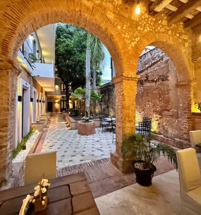 "Patio interior visto a través de un arco de piedra iluminado, que combina un muro antiguo de ladrillo a la derecha, un edificio moderno a la izquierda y un jardín central con palmeras y baldosas decorativas."