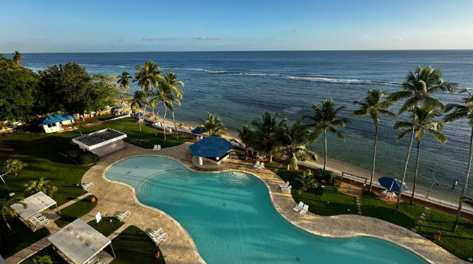 Piscina exterior de un resort frente al mar con palmeras, un gazebo de techo azul y una vista panorámica del océano en un día soleado