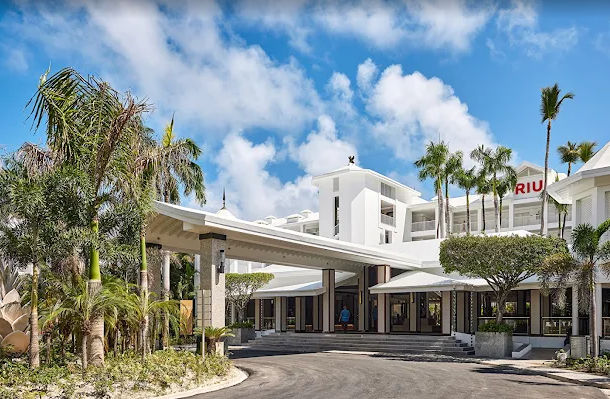 Main entrance of the RIU Hotel with a white facade and palm trees under a blue sky.