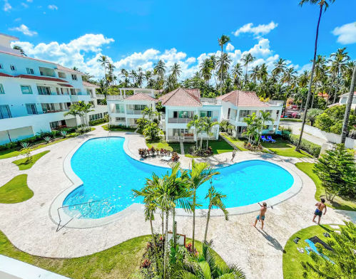 Panoramic view of an irregularly shaped swimming pool surrounded by white villas with tiled roofs, palm trees and landscaped areas in a Punta Cana resort.