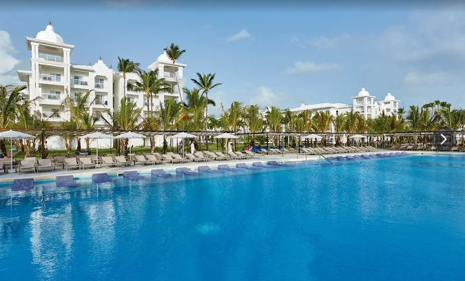"Piscina cristalina rodeada de palmeras frente a las modernas instalaciones del resort en Punta Cana, ideal para disfrutar de unas vacaciones de lujo tras un cómodo traslado privado con Taxi Dominicana."