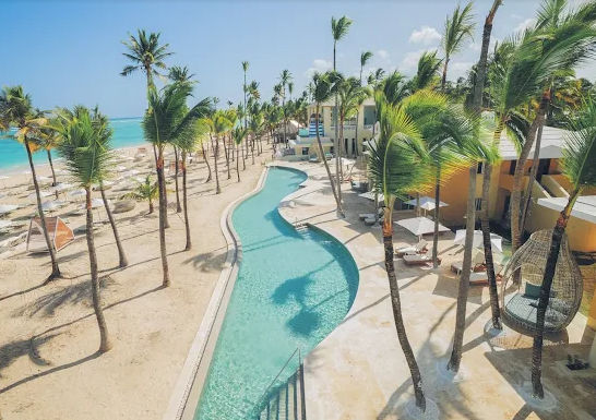Vista aérea de una piscina sinuosa que bordea una playa de arena blanca con palmeras y tumbonas frente al mar Caribe en un resort de Punta Cana.