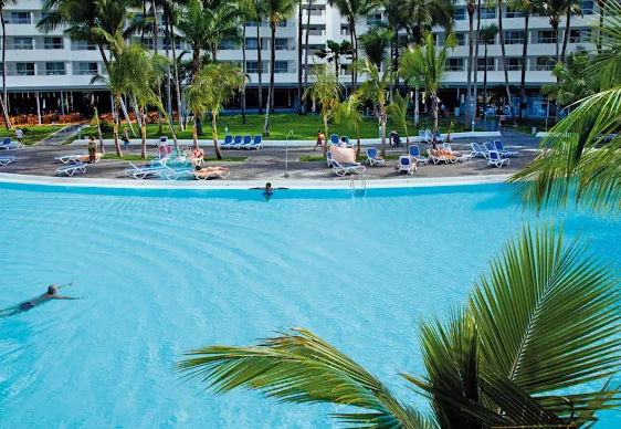 "View of a large swimming pool with crystal clear blue water at a hotel complex, with people swimming, sun loungers and palm trees in the relaxation area in front of a hotel building."