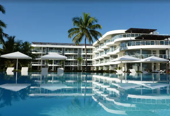 "Panoramic view of a modern infinity pool in front of a luxury resort with curved architecture, surrounded by palm trees under a clear sky."