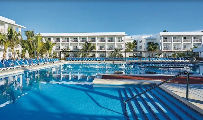 "Vista panorámica de la piscina principal de un resort en Punta Cana, con aguas cristalinas, escalinatas de acceso, numerosas hamacas dispuestas para los huéspedes y el moderno edificio del complejo hotelero de fondo bajo un cielo despejado."