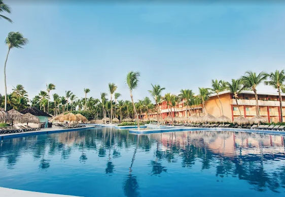  View of a large crystal clear pool at a resort in Punta Cana, surrounded by palm trees and thatched umbrellas under a clear sky.