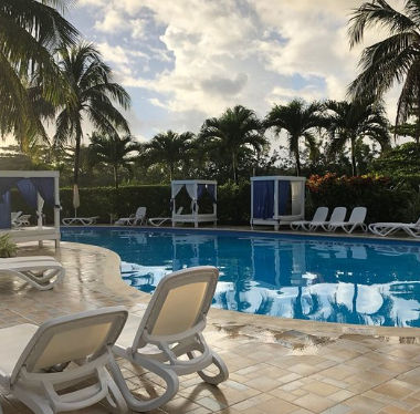 An outdoor swimming pool surrounded by palm trees under a partly cloudy sky. In the foreground are several white sun loungers on a stone patio. Around the pool are additional sun loungers and cabanas covered with blue and white fabrics.