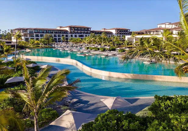"Vista panorámica de una piscina de diseño moderno y curvilíneo rodeada de exuberante vegetación tropical, palmeras y sombrillas, con un complejo hotelero de estilo elegante al fondo bajo un cielo despejado."