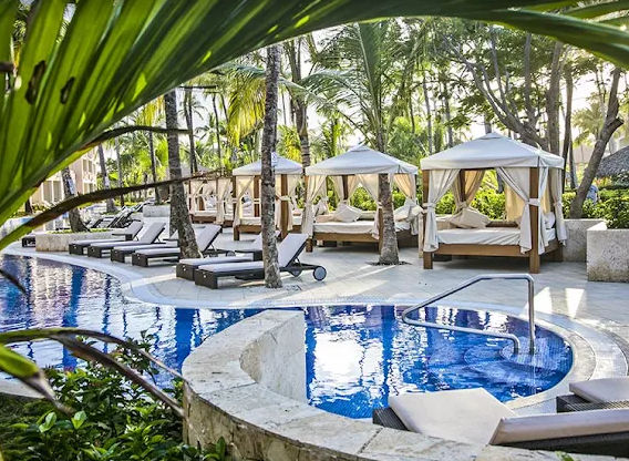 "Luxury view by a blue-tiled pool at a resort, highlighting several Balinese beds with white curtains, sun loungers and palm trees in a tropical setting."